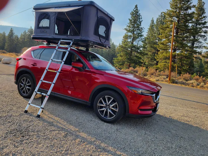 Red Mazda CX-5 with a rooftop tent & ladder, parked on gravel. A versatile RV rental for nature camping.. Mazda Campervan 2018