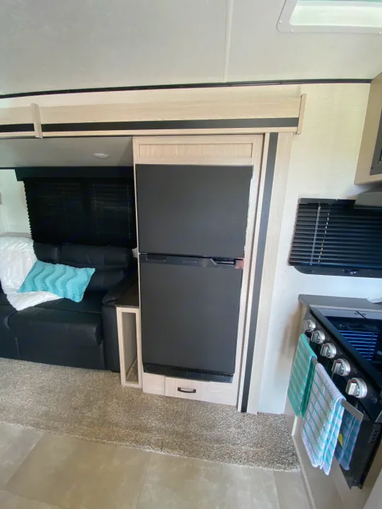 Inside an RV rental: a sleek refrigerator next to a cozy black couch, with a compact kitchen area nearby.. Coachmen Other 2021