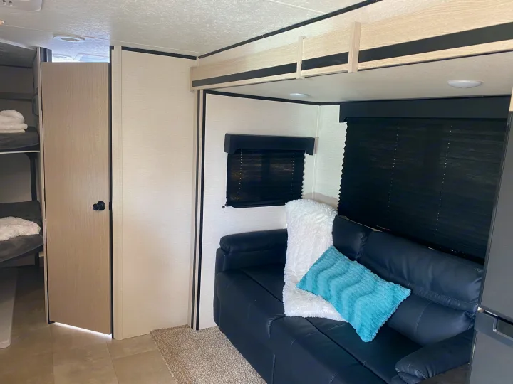 Modern RV rental interior: black sofa with accent pillows, privacy blinds, and a glimpse of bunk beds through an open doorway.. Coachmen Other 2021