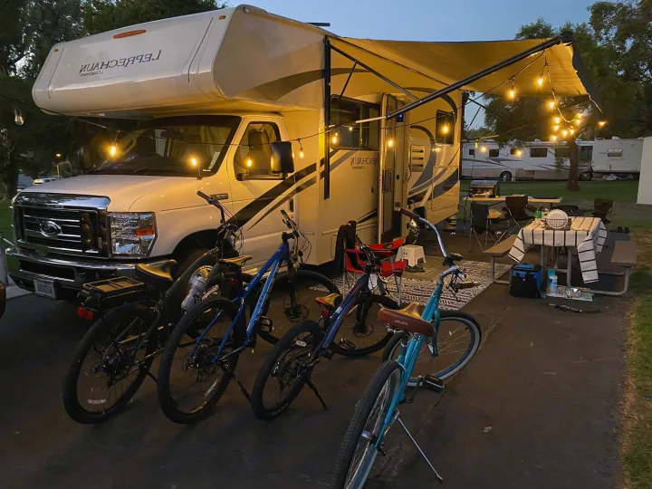An RV rental illuminates a cozy campsite at dusk with string lights. Bicycles and a picnic area complete this inviting outdoor scene.. Coachmen Leprechaun 2021