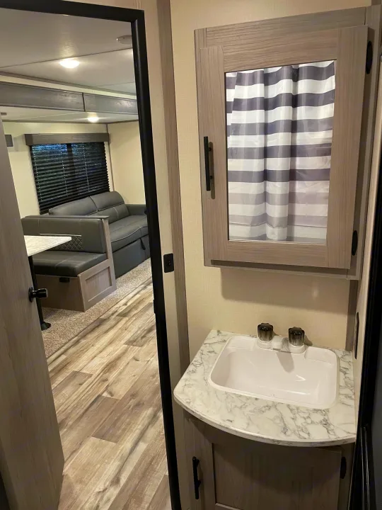 Clean RV rental bathroom vanity with sink and mirrored cabinet. Reflection shows a cozy living area with gray sofa.. Dutchmen Coleman 2022