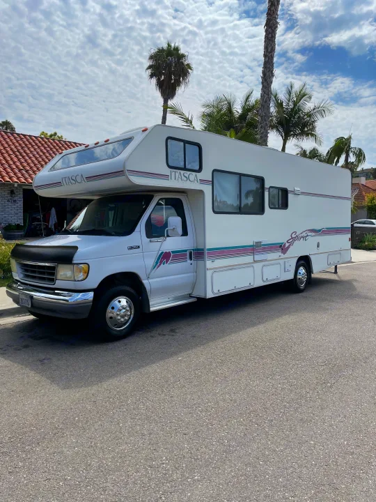 White Itasca Spirit Class C RV rental parked on a sunny street with palm trees and a tiled-roof house.. Itasca Spirit 1995