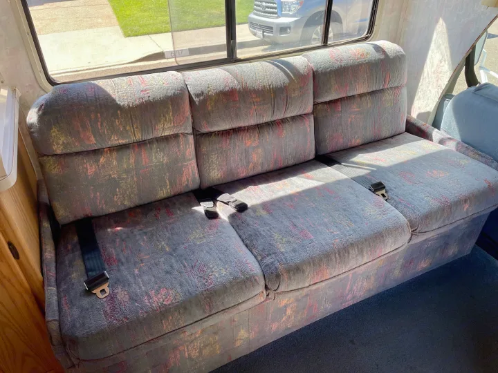 RV rental interior: Long, patterned sofa with three seatbelts, sunlit through the window, offering passenger comfort.. Itasca Spirit 1995