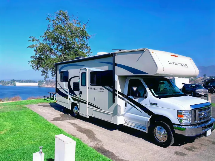 A Class C RV rental parked at a scenic campsite overlooking a lake and dam, with mountains beyond.. Coachmen Leprechaun 2021
