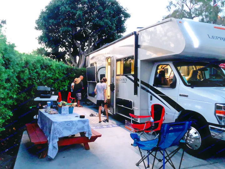 Family enjoying an RV rental. Evening outdoor setup with picnic table, chairs, and illuminated RV.. Coachmen Leprechaun 2021