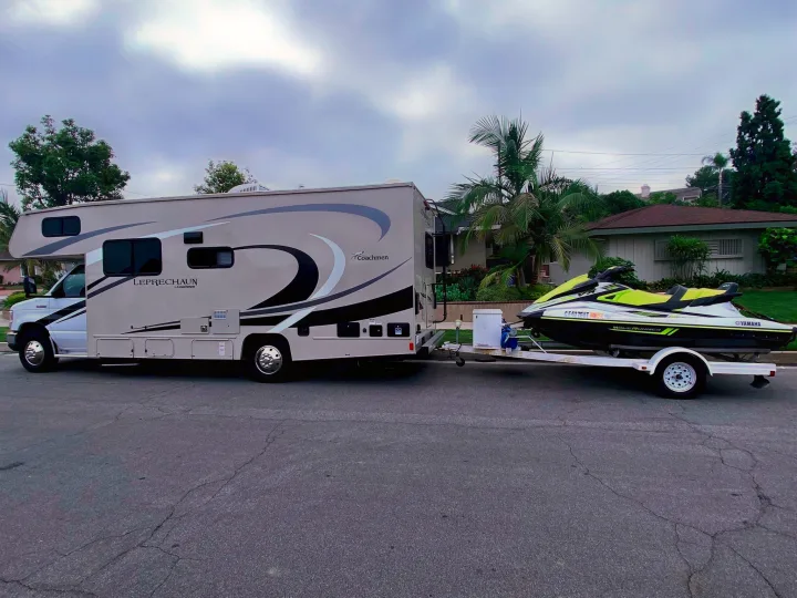 RV rental towing a lime green Yamaha WaveRunner jet ski on a trailer, parked on a street, ready for outdoor travel.. Coachmen Leprechaun 2021