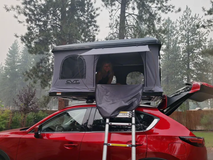 Woman smiles from rooftop tent on red SUV. Perfect RV rental for elevated camping adventures amidst hazy forest pines.. Mazda Campervan 2018