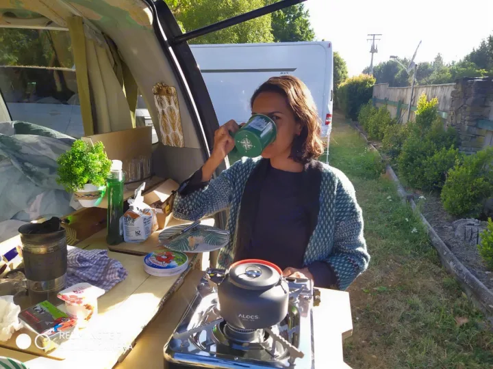 A woman sips from a green mug next to her RV rental, with a portable stove and breakfast setup.. Toyota Noah 2005