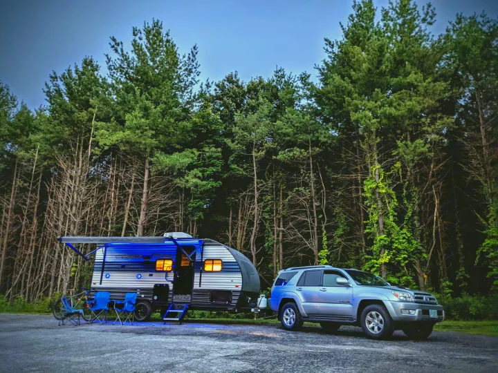 An RV rental travel trailer, illuminated by blue lights, and an SUV are parked at a serene wooded campsite at dusk.. Forest River Cherokee Wolf Pup 2021