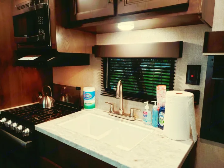 Compact RV rental kitchen with stove, microwave, sink, and cleaning supplies on the countertop, ready for use.. Jayco Jay Flight SLX 2021