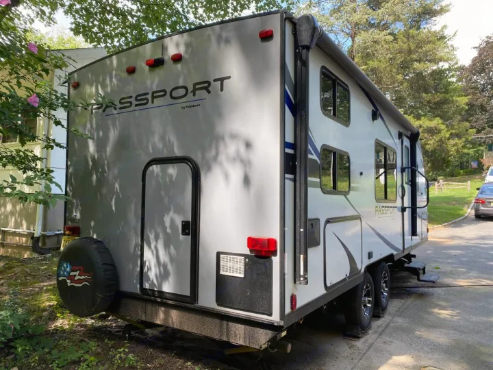 A white Passport travel trailer, a type of RV rental, is parked outside a house with trees. Shows rear and side.. Other Passport 2020