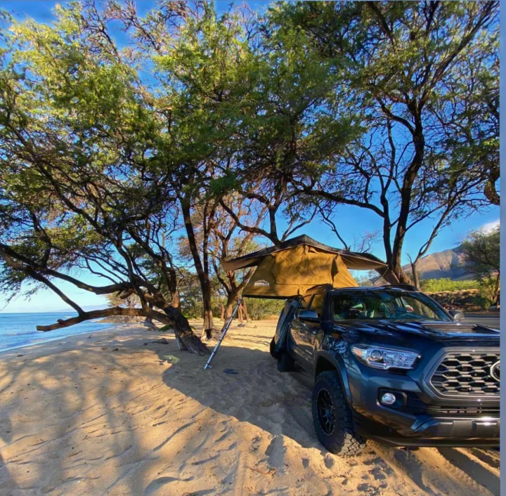 A pickup truck with an open rooftop tent is parked for beach camping under trees, a unique RV rental experience.. toyota tacoma 2021