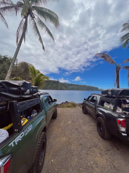 Two overland trucks with rooftop tents, like an RV rental setup, by a scenic bay with mountains & palms. Perfect for adventure!. Toyota Tacoma 2021