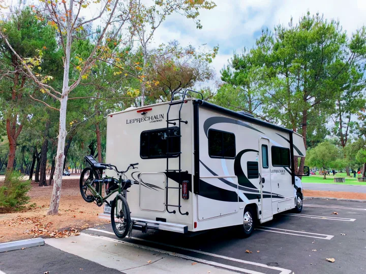 A beige LEPRECHAUN RV rental with two bicycles on a rear rack is parked in a tree-lined lot at a park.. Coachmen Leprechaun 2021