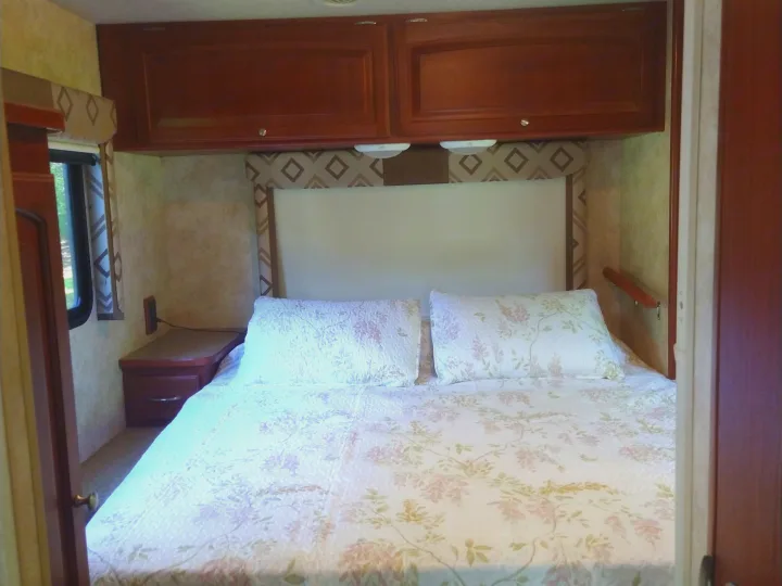 Cozy RV rental bedroom with a comfortable bed, overhead cabinets, and a nightstand, perfect for sleeping on your trip.. Winnebago Access 2011