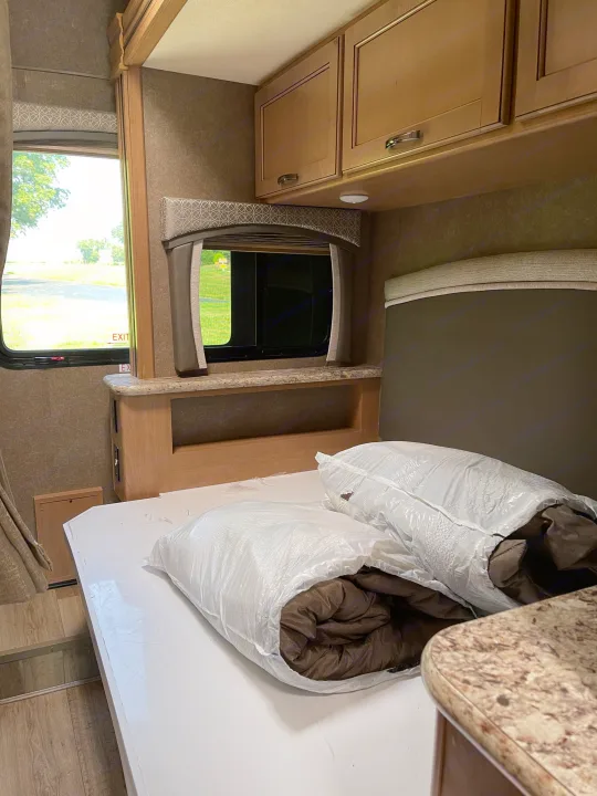 RV rental bedroom area: A neatly made bed, still wrapped in plastic, with overhead cabinets, a window, and wood flooring.. THOR FOUR WINDS 26 B 2021