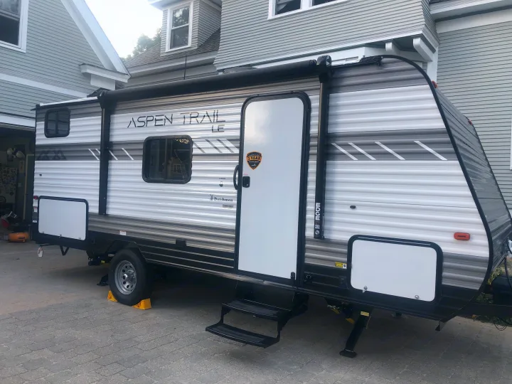 A clean Aspen Trail LE travel trailer, perfect for an rv rental, is parked on grey pavers next to a house, stabilized and ready.. Dutchman Aspen Trail 2022