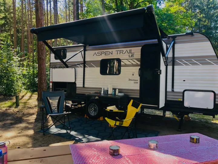 RV rental at a wooded campsite with an extended awning, outdoor chairs, table, and a picnic table in the foreground.. Dutchman Aspen Trail 2022