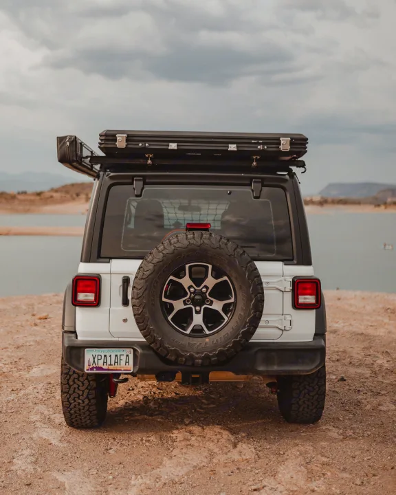A white Jeep Wrangler with a rooftop tent, ideal for an RV rental adventure, is parked by a scenic lake under cloudy skies.. Jeep Wrangler Unlimited 2020