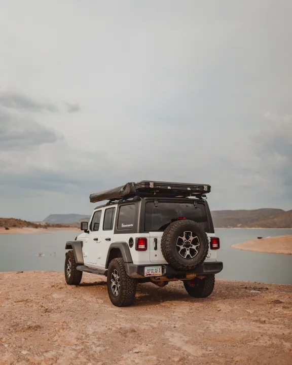 Adventure-ready white Jeep Wrangler RV rental with rooftop tent by a scenic lake, perfect for overlanding excursions.. Jeep Wrangler Unlimited 2020