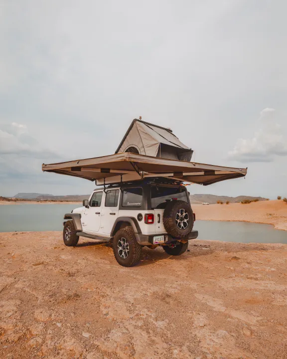 White Jeep Wrangler with rooftop tent & awning by a lake, ideal for an RV rental outdoor escape.. Jeep Wrangler Unlimited 2020