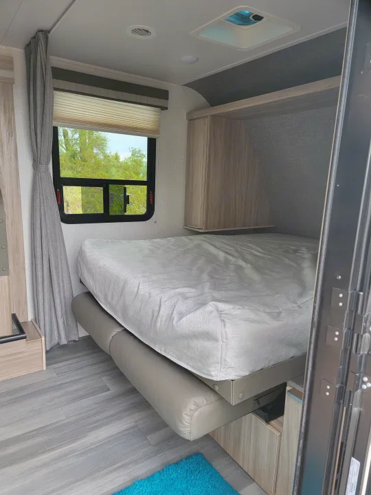 RV rental bedroom with a light gray bed, wooden overhead cabinets, and a window displaying an 'EXIT' sign. Features sleek, gray flooring.. Winnebago Micro Minnie 2021