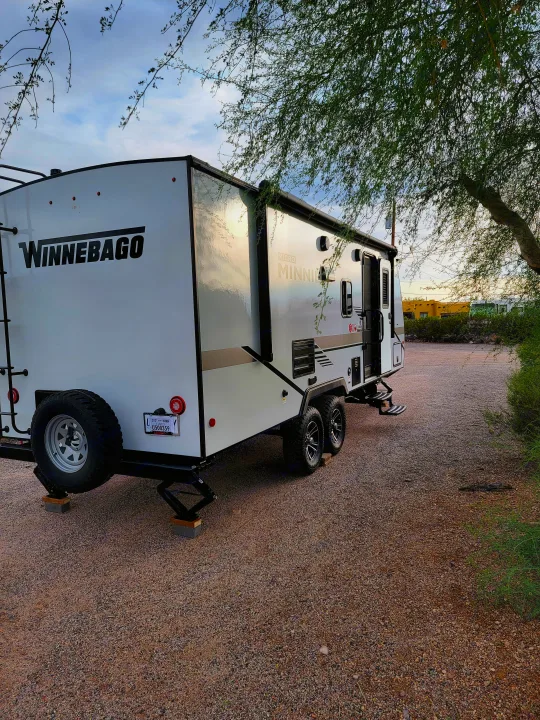 White Winnebago Micro Minnie RV rental parked on a gravel lot, stabilizers down, under a tree canopy. Perfect outdoor stay.. Winnebago Micro Minnie 2021