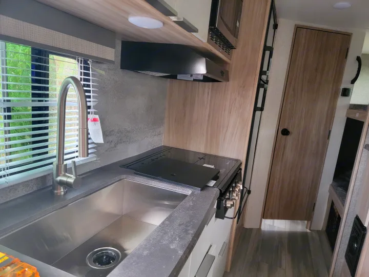 Modern RV rental kitchen featuring a stainless steel sink, pull-down faucet, counter, cooktop, and microwave. Compact and functional space.. Winnebago Micro Minnie 2021