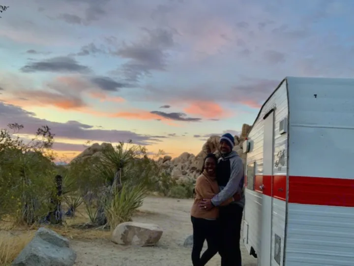 A couple hugs by their RV rental in a scenic desert landscape at sunset, surrounded by rocks and yuccas.. Shasta Compact 1968
