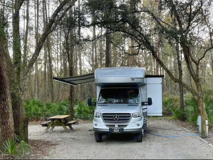 Silver RV rental parked at a serene campsite with an extended awning and slide-out, surrounded by trees and a picnic table.. Winnebago View 2022