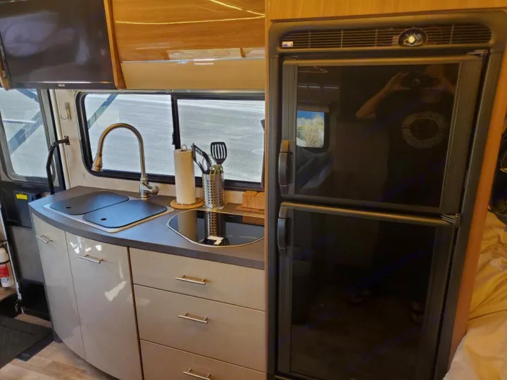 Interior of an RV rental: a modern kitchen with a dual-basin sink, induction cooktop, cabinets, and a tall, black two-door refrigerator.. Winnebago View 2018