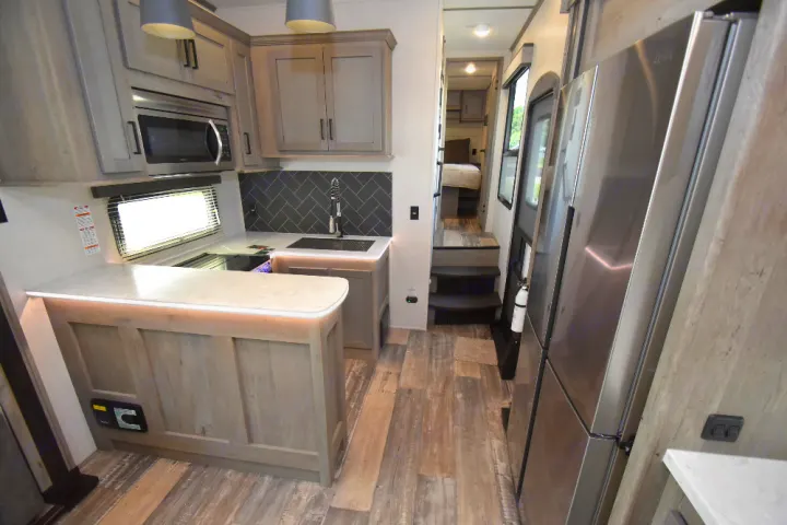 Modern RV rental interior: kitchen with light wood cabinets, stainless appliances, island, and steps to a raised bedroom.. Keystone Raptor 2025