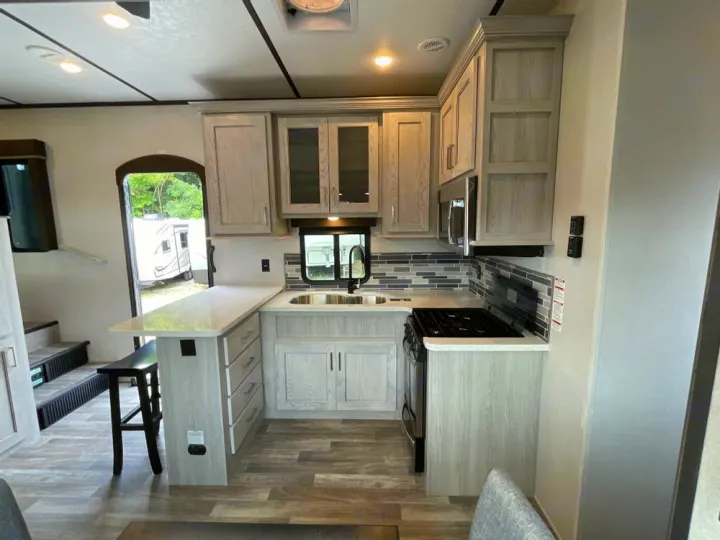 Step inside this modern RV kitchen, featuring light cabinets, white counters, and a full stove, ideal for your RV rental experience.. Keystone RV Montana High Country 2024