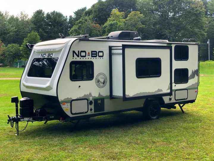 This light-colored No Boundaries travel trailer with an extended slide-out and roof rack is parked on green grass, ideal for RV rental.. No Boundaries 19 2021