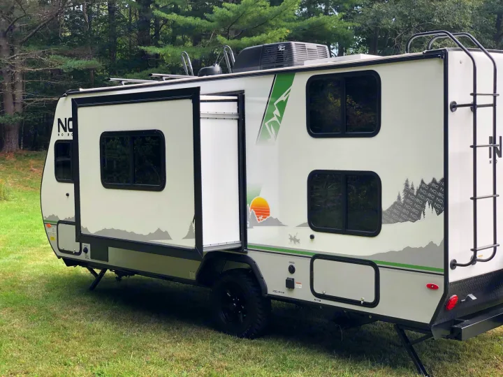A light-colored travel trailer with an open side door, rooftop AC unit, and graphics, ready for an rv rental adventure in nature.. No Boundaries 19 2021