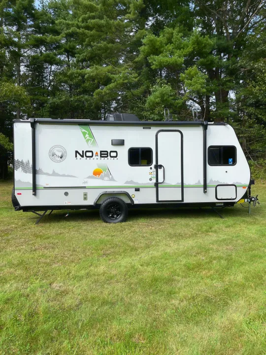 White No Boundaries travel trailer RV rental, seen parked on grass with a forest backdrop. Perfect for your next outdoor trip.. No Boundaries 19 2021