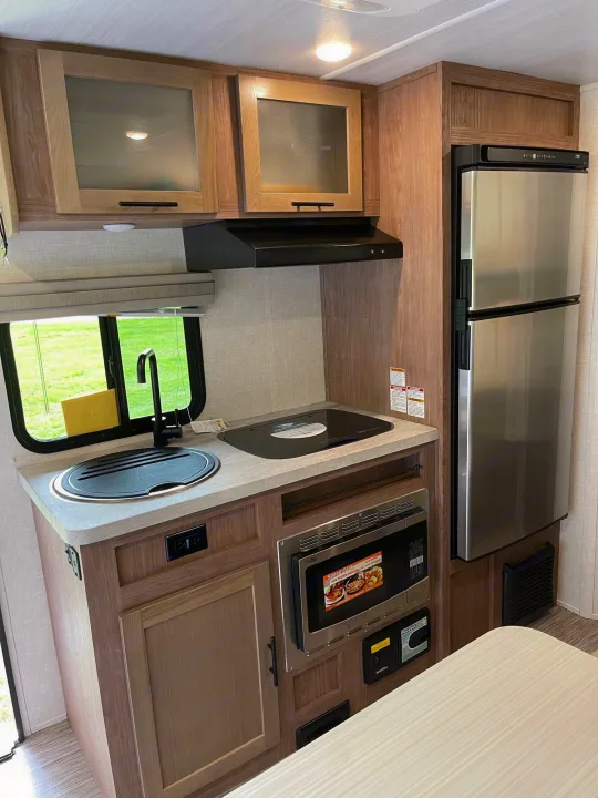 An rv rental's modern kitchen features a sink, stovetop, stainless fridge, microwave, and light wood cabinetry.. No Boundaries 19 2021