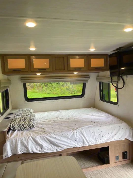 Interior of an RV rental bedroom showing a bed, overhead cabinets, and windows with green outdoor views.. No Boundaries 19 2021