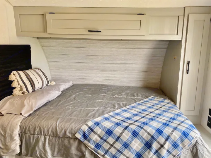 Cozy RV rental bedroom with light wood interior, overhead cabinets, and a neatly made bed with plaid blanket.. Forest River Wildwood 2021