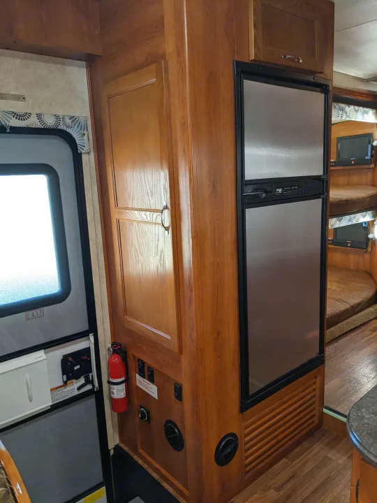 RV rental interior with wooden cabinets, large refrigerator, and bunks. Exit door and fire extinguisher are near controls.. Forest River Coachmen Freelander 2011
