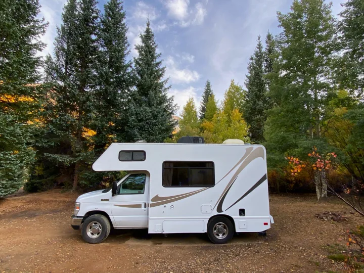 White RV rental parked on a dirt road, amidst towering evergreen and vibrant autumn trees under a cloudy sky.. Thor Motor Coach Four Winds Majestic 2017