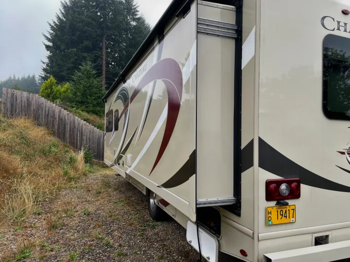 A beige "Chateau" RV rental, with its slide-out extended, parked on gravel beside a wooden fence and trees, ready for travel.. Thor Motor Coach Chateau 2017