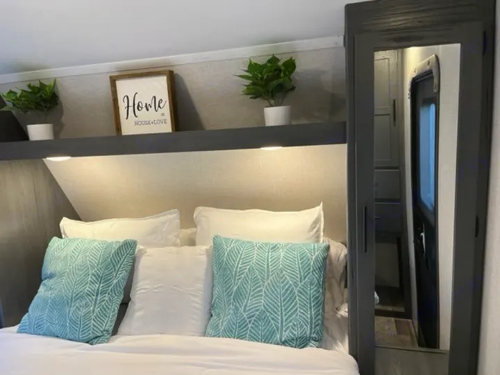 This cozy RV rental bedroom features a bed with teal and white pillows, decorative shelf, and mirrored door.. Dutchmen Aspen Trail 2021