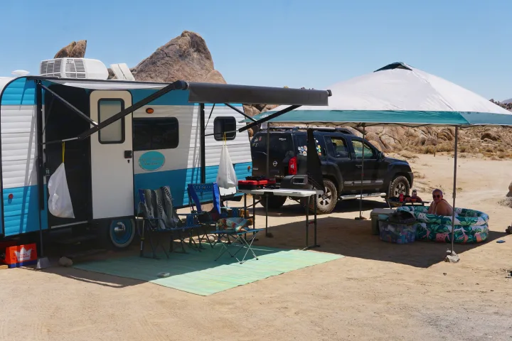 Desert RV rental setup: blue and white travel trailer, SUV, shade canopy, and a person relaxing in a small pool. Ideal for outdoor adventures.. Old School 818 2021