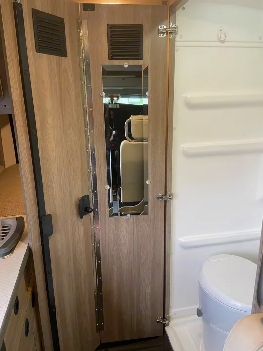 Compact RV rental bathroom featuring a wood door with an integrated mirror, and a white toilet inside.. Winnebago Solis 2021