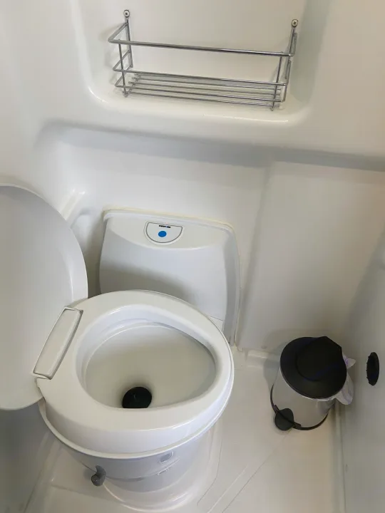 This compact RV rental bathroom features a white toilet, an overhead wire shelf, and a small waste bin on the floor.. Winnebago Solis 2021