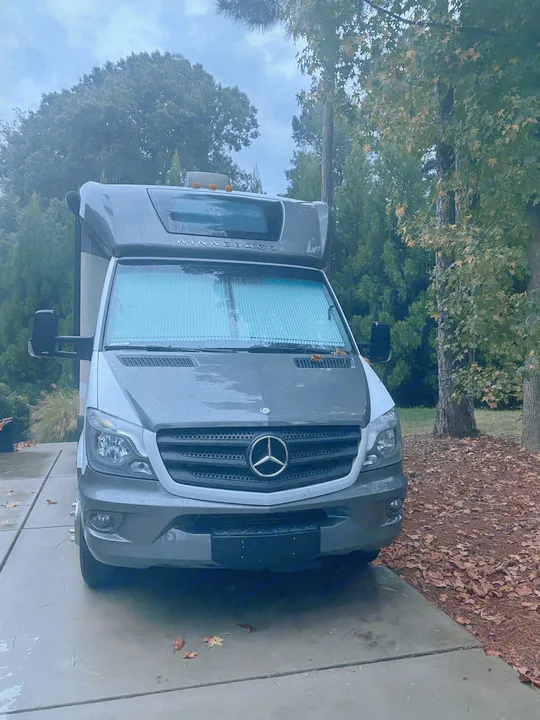 Grey/white Mercedes Sprinter RV rental parked on a wet driveway surrounded by trees and autumn leaves on an overcast day.. Winnebago View 2016
