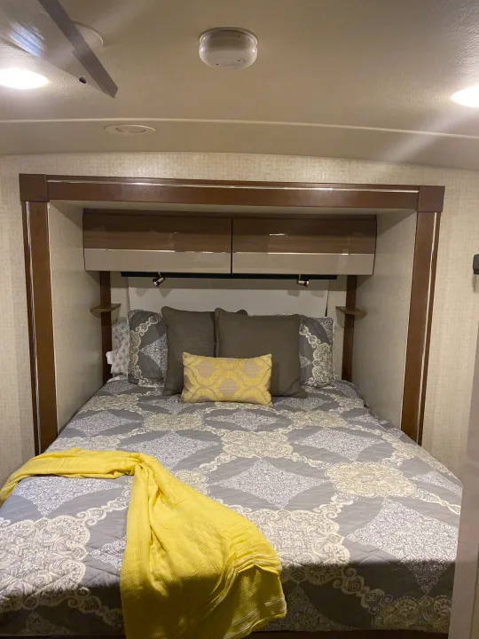 Cozy RV rental bedroom featuring patterned bedding, a yellow throw, overhead storage, and reading lights for a comfortable stay.. Winnebago View 2016