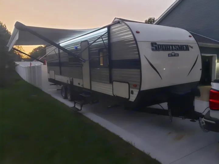 This Sportsmen SE travel trailer is ready for your RV rental adventure. Awning extended, parked on a driveway at dusk.. K-Z Manufacturing Sportsmen 2021