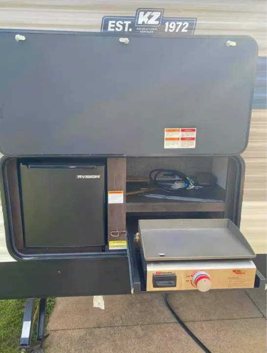 An open RV rental compartment reveals a compact fridge & pull-out griddle, part of a convenient outdoor kitchen.. K-Z Manufacturing Sportsmen 2021